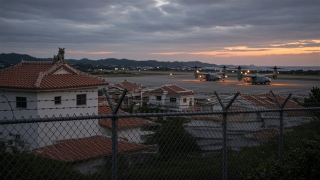 Okinawan houses with red-tiled roofs and a shisa lion statue beside a military airfield with tiltrotor aircraft, seen through a barbed-wire fence at dusk